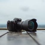 A DSLR camera with a telephoto lens is positioned on a wooden surface against a blurred waterscape background.