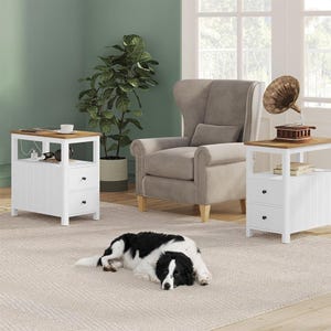 A white side table featuring a single drawer, lower shelf, and an integrated power outlet on its top surface, positioned next to an upholstered armchair.