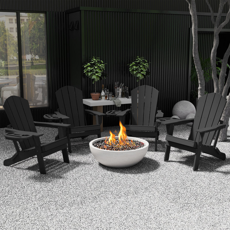 Four black Adirondack chairs surround a round fire pit filled with stones. A nearby table holds various beverages, all set in a modern patio area with plants and textured surfaces.