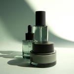 Three skincare containers: a dropper bottle, a tall lotion bottle, and a round jar, all with dark lids and clear glass.