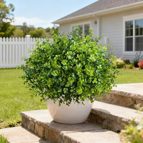 A round artificial boxwood shrub in a white pot is placed outdoors on stone steps, with a house and garden in the background.