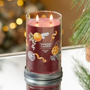 A Yankee Candle with the scent \“Holiday Zest,\“ featuring a festive design with oranges, pine branches, and ribbon elements, is illuminated with two lit wicks, against a blurred holiday background.
