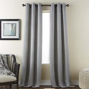 Gray curtains with a subtle checkered pattern hang from a black rod in front of a window. To the left is a chair with a textured throw, and a framed abstract painting hangs on the wall.
