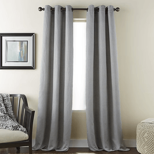 Gray curtains with a subtle checkered pattern hang from a black rod in front of a window. To the left is a chair with a textured throw, and a framed abstract painting hangs on the wall.