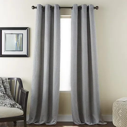 Gray curtains with a subtle checkered pattern hang from a black rod in front of a window. To the left is a chair with a textured throw, and a framed abstract painting hangs on the wall.