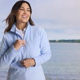 A woman wears a light blue fleece pullover with a white collared shirt underneath, standing near a body of water with a landscape in the background.