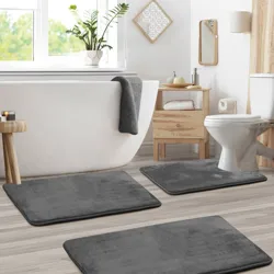 Bathroom scene featuring a white freestanding bathtub, a toilet, a wooden storage unit, and dark gray bath mats. Accessories include a plant, towels, and toiletries on a wooden tray.