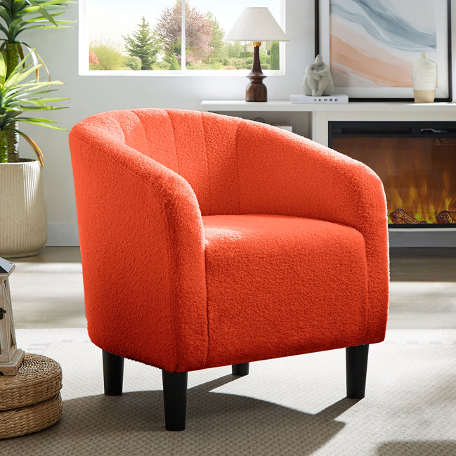 An orange teddy barrel chair with a textured fabric, featuring a rounded back and armrests. It has black legs and is positioned in a cozy living room setting near a window, with a decorative plant and a fireplace in the background.