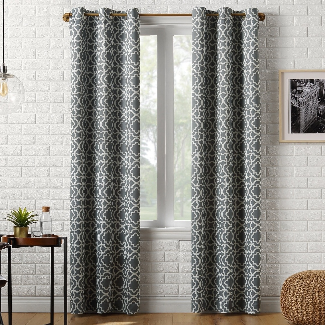 Gray curtains with a white geometric pattern hang on a wooden rod in front of a window. The setting includes a white brick wall, a framed black-and-white photo, a small table with a plant and glass, and a woven floor pouf.