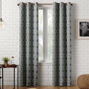 Gray curtains with a white geometric pattern hang on a wooden rod in front of a window. The setting includes a white brick wall, a framed black-and-white photo, a small table with a plant and glass, and a woven floor pouf.