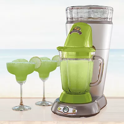A green Margaritaville drink maker with a clear ice reservoir on top and a blending jar below, next to two margarita glasses filled with a frozen green beverage on a beach backdrop.