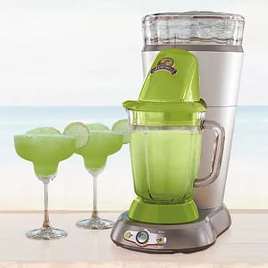 A green Margaritaville drink maker with a clear ice reservoir on top and a blending jar below, next to two margarita glasses filled with a frozen green beverage on a beach backdrop.