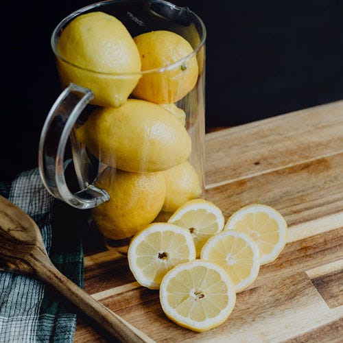 lemon slices beside a clear glass pitcher with lemon stacked inside on top of a wooden cutting board