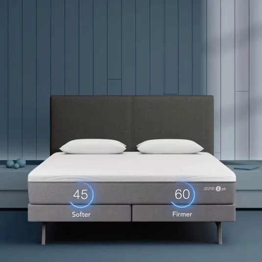 A bed with an adjustable mattress showing dual firmness levels of 45 (softer) and 60 (firmer), accompanied by two pillows; set in a modern bedroom with a blue paneled wall.