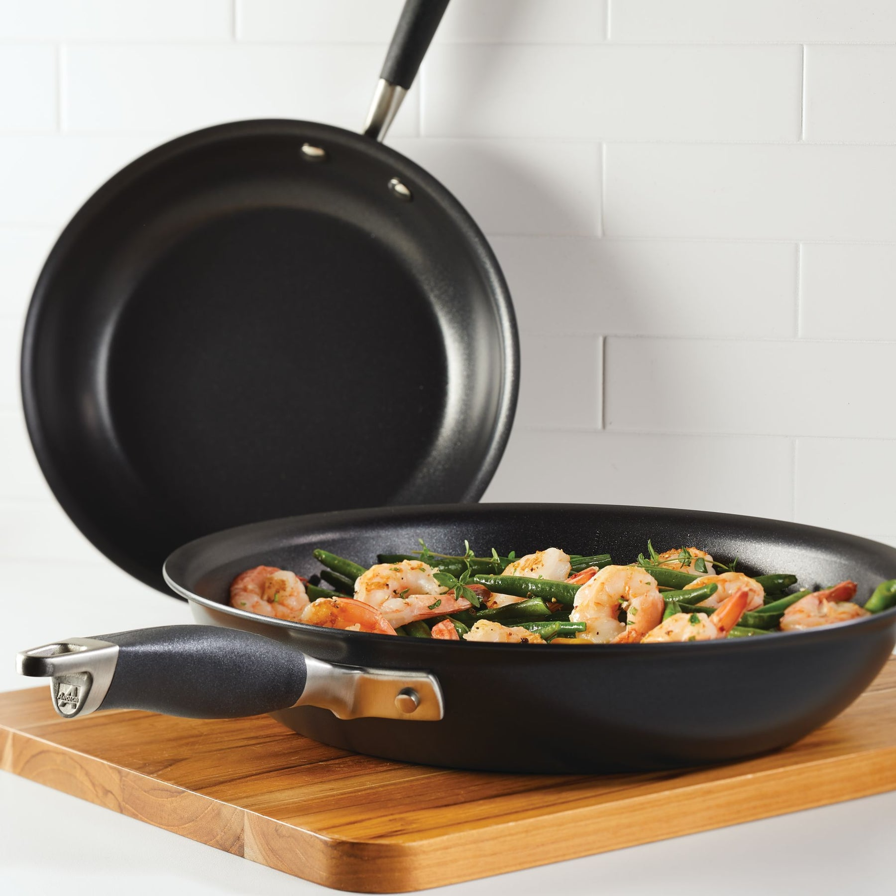 Two non-stick frying pans on a wooden board, one with sautéed shrimp and green beans.