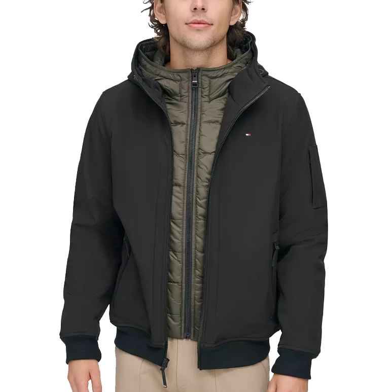 A man is wearing a black and khaki Tommy Hilfiger hooded bomber jacket with a zip-up front, side pockets, and logo on the arm.