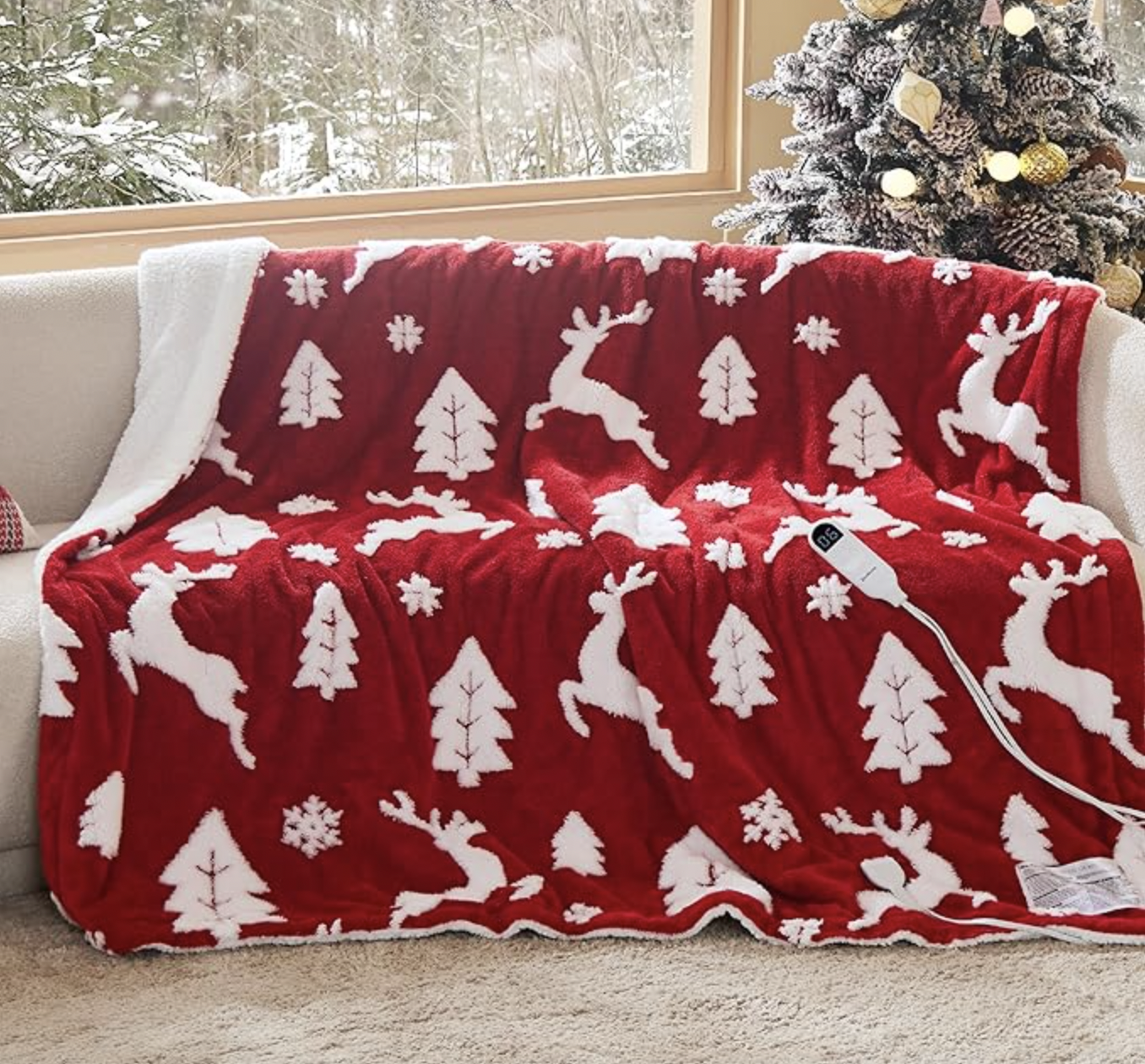 A red and white festive blanket with reindeer, trees, and snowflakes, draped over a sofa near a decorated, snow-covered Christmas tree.