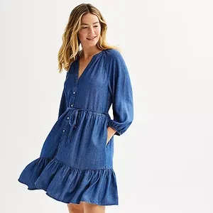A woman is wearing a long-sleeved denim dress with a button front, a drawstring waist, and a ruffled hem.
