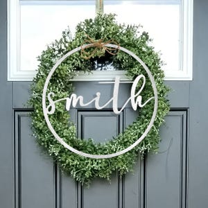 A circular wreath made of green foliage, decorated with a wooden ring featuring the word \“smith\“ in cursive, hangs on a door.