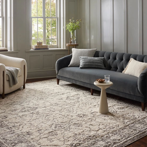 Living room with a dark gray tufted sofa, a light beige armchair, a textured cream and gray area rug, a small round side table, and a few pillows. Large windows and wood panel walls complete the space.