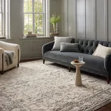 Living room with a dark gray tufted sofa, a light beige armchair, a textured cream and gray area rug, a small round side table, and a few pillows. Large windows and wood panel walls complete the space.