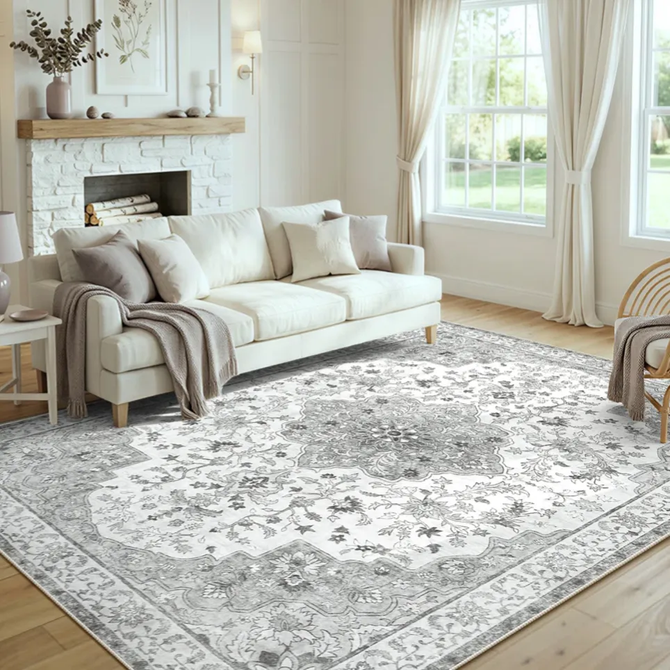 A cozy living room featuring a large, ornate area rug with a floral pattern, a white sofa with neutral cushions and throws, wicker chairs, a fireplace with a wooden mantel, and a ceiling fan with wooden blades.