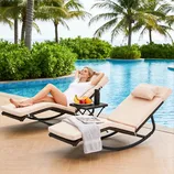 Two beige cushioned lounge chairs with black metal frames are by a pool, accompanied by a small table holding a towel, magazine, fruit, and a thermos.