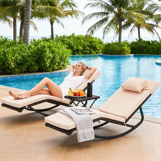 Two beige cushioned lounge chairs with black metal frames are by a pool, accompanied by a small table holding a towel, magazine, fruit, and a thermos.