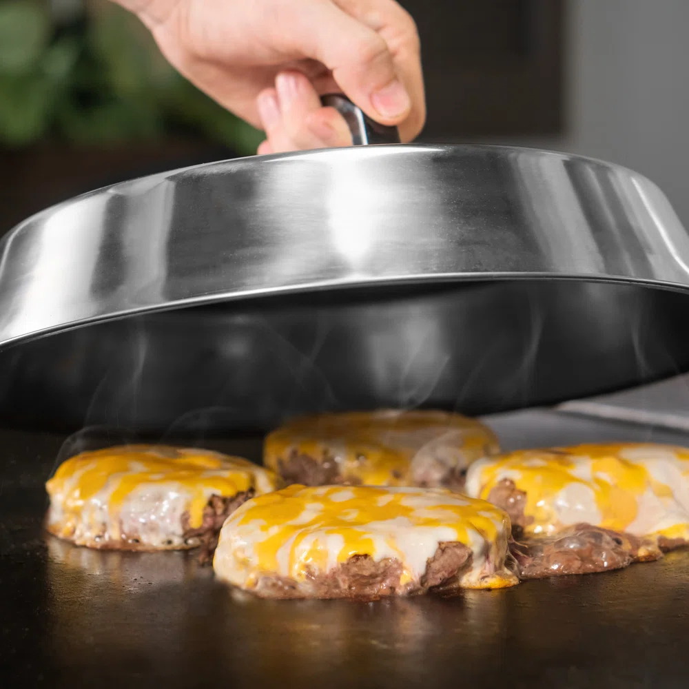 Cheese-topped hamburger patties cooking on a griddle, with a hand lifting a stainless steel dome cover.