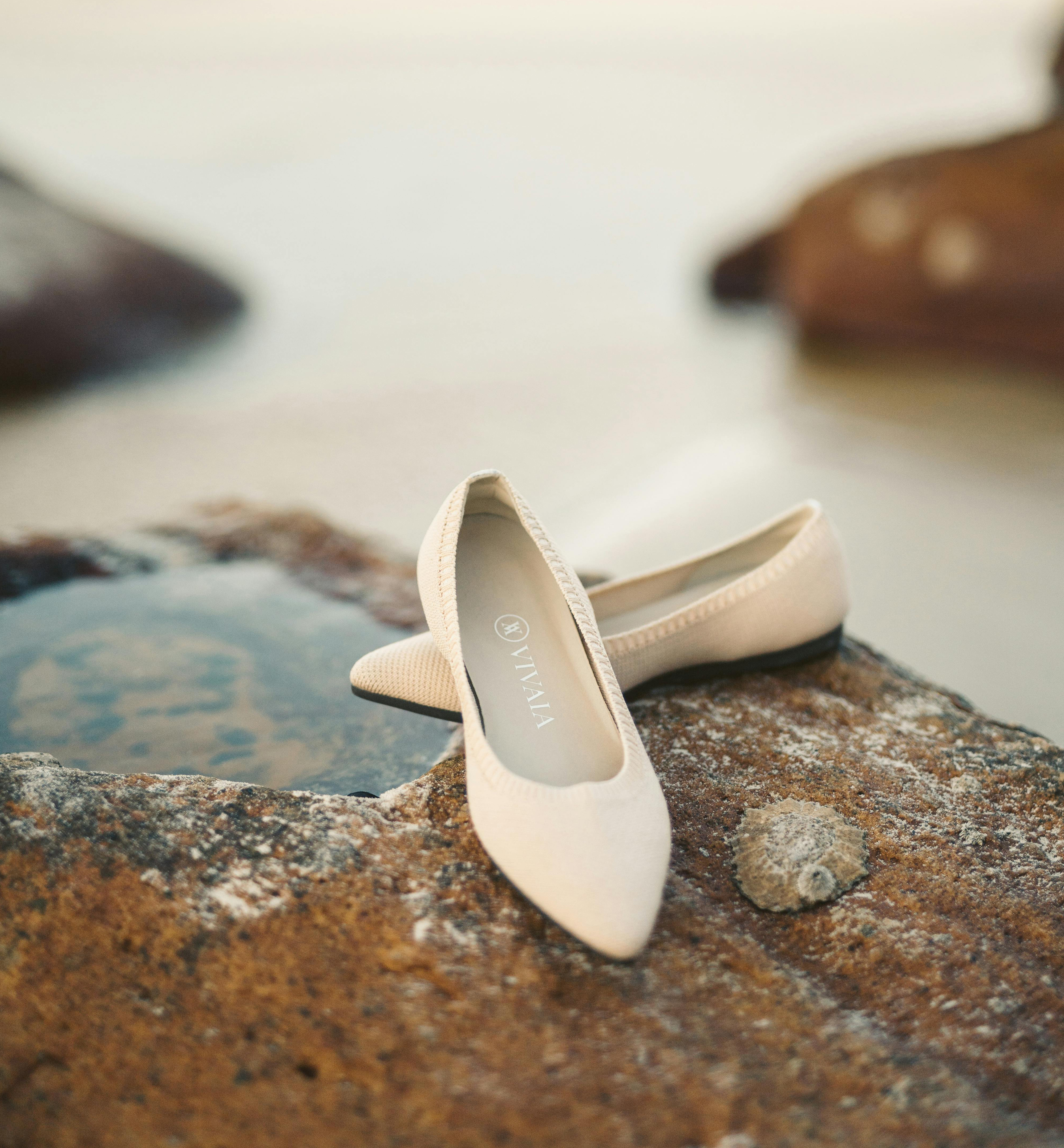 A pair of beige pointed-toe flats resting on a rock by water.