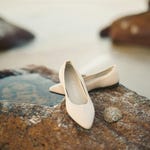 A pair of beige pointed-toe flats resting on a rock by water.