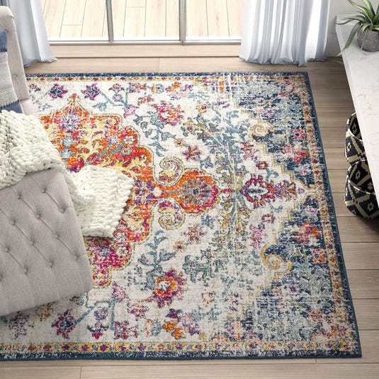 Colorful area rug with a vibrant, intricate floral pattern in orange, pink, blue, and yellow tones, placed in a living room setting with a gray tufted sofa and a chunky knit blanket.