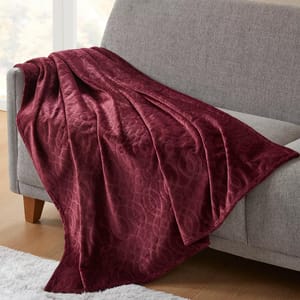 A maroon, textured throw blanket with an intricate pattern is draped over a light gray couch, next to a white rug.