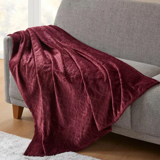 A maroon, textured throw blanket with an intricate pattern is draped over a light gray couch, next to a white rug.