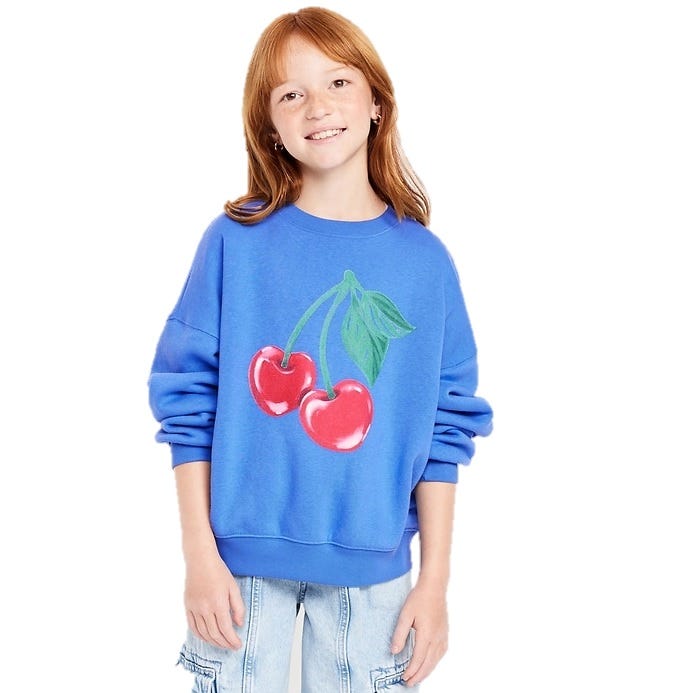 A young person wears a blue sweatshirt with a large cherry graphic on the front, paired with light-wash jeans.