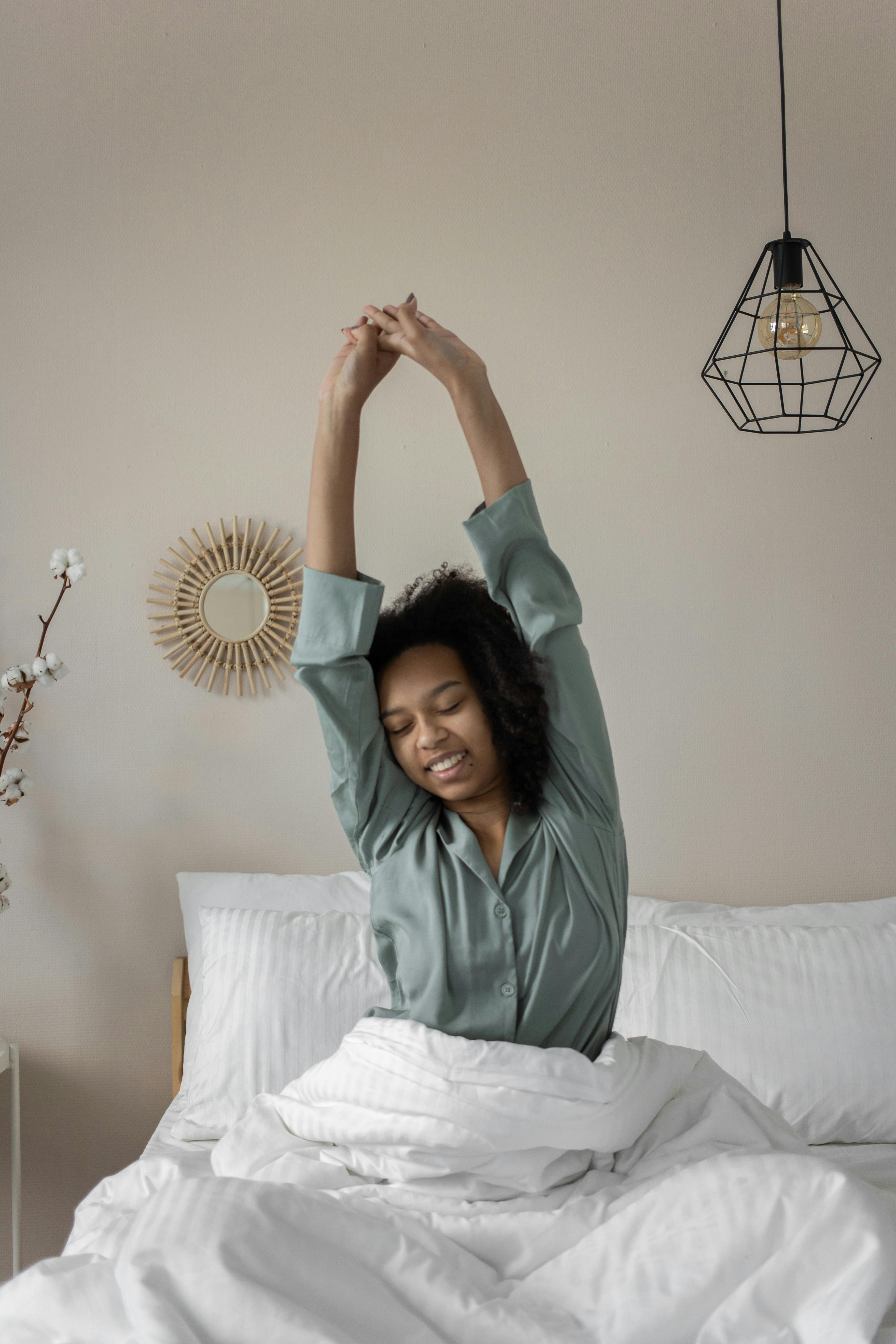 A woman stretching in bed with white bedding, beside a hanging geometric lamp and a round mirror on the wall.