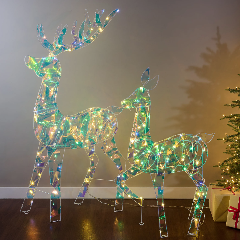 Two illuminated reindeer figures with LED lights are depicted, featuring a holographic design. A decorated Christmas tree and a wrapped gift box are nearby, creating a festive atmosphere.