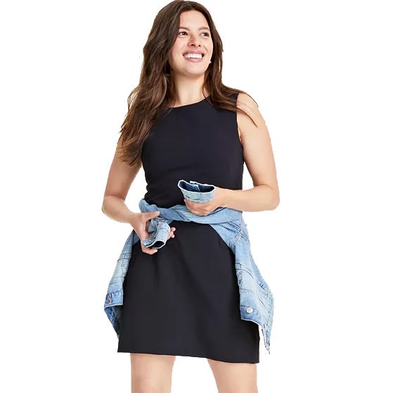 A woman is wearing a black sleeveless mini dress with a fitted bodice and an A-line skirt, paired with a blue denim jacket tied around her waist.