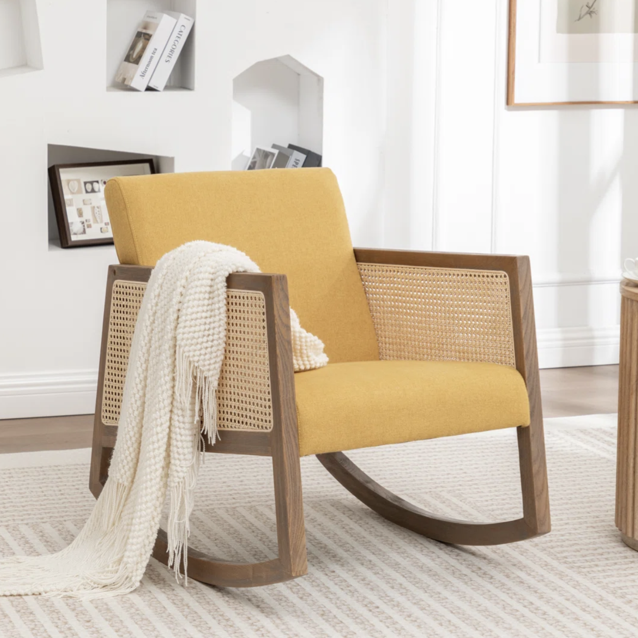 Yellow cushioned rocking chair with wooden frame, featuring a woven caned design on the sides, draped with a white knit throw. Placed on a textured cream and beige rug, next to a round wooden side table with a teacup.