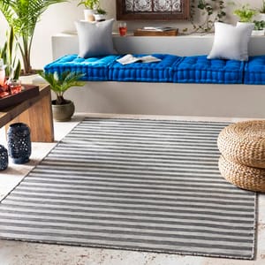 A cozy living space features a blue cushioned bench with pillows, a striped rug, woven poufs, a wooden table with lanterns, and various potted plants.
