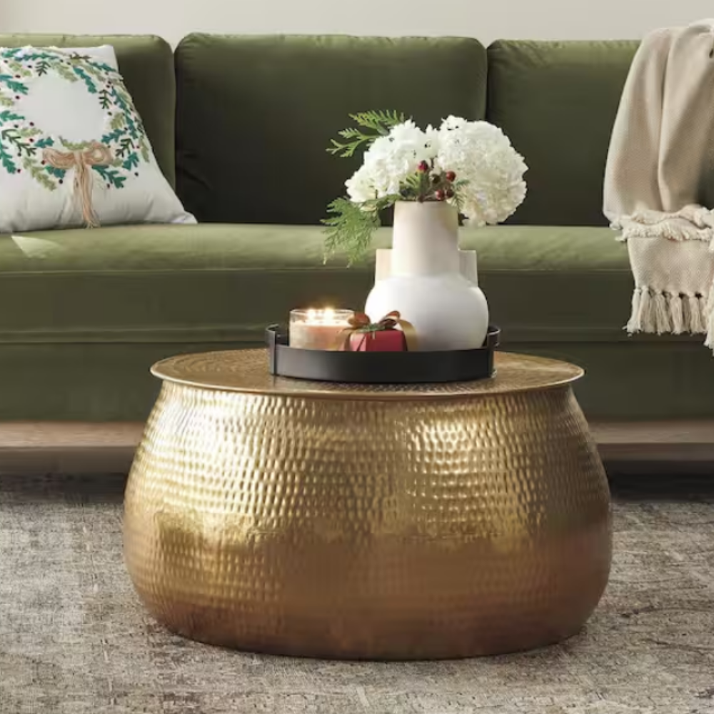 A round, gold, hammered metal coffee table is topped with a black tray holding a white vase with flowers, candles, and a small gift. The table sits on a rug, with a green sofa and decorative pillow in the background.