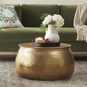 A round, gold, hammered metal coffee table is topped with a black tray holding a white vase with flowers, candles, and a small gift. The table sits on a rug, with a green sofa and decorative pillow in the background.