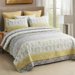 A bedding set featuring a quilt and shams with a floral pattern in yellow, gray, and white tones on a bed with a padded headboard.