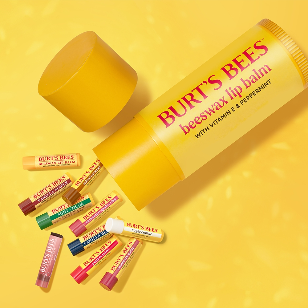 Burt's Bees lip balms in various flavors, including beeswax with vitamin E and peppermint, vanilla maple, mint cocoa, and sugar cookie, are displayed against a yellow background.