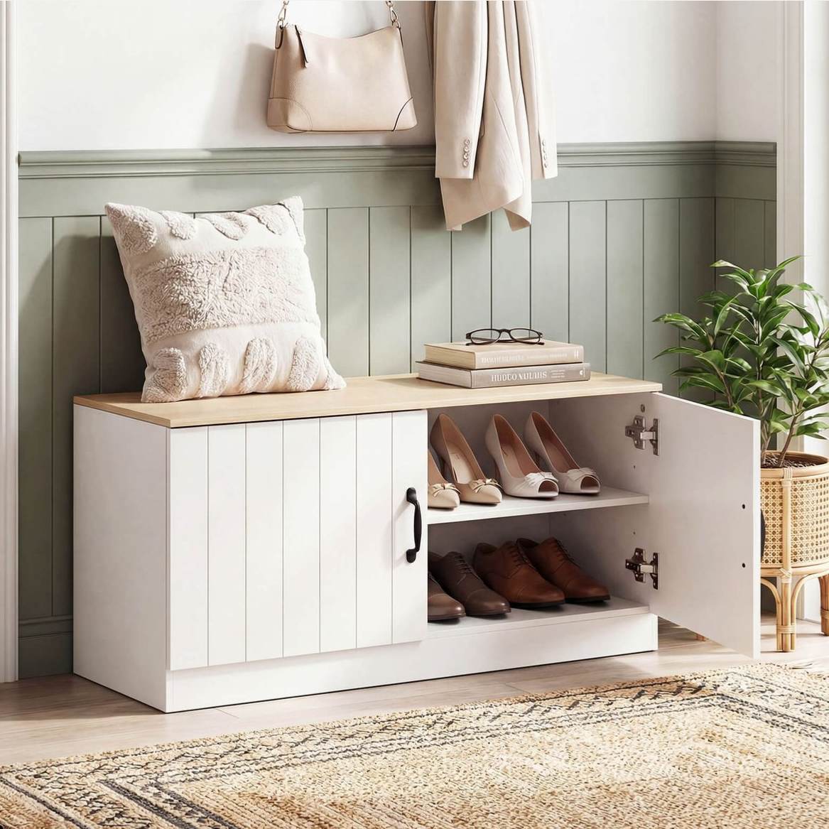 A cozy entryway features a white storage bench with a wooden top, holding a textured pillow and books. A cabinet with shoes is open, while a coat, bag, and potted plant accessorize the space.