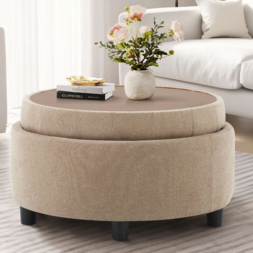 A round, beige ottoman with a fabric texture and dark legs doubles as a coffee table, featuring a tray with a vase of flowers and stacked books.