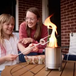 A small, portable fire pit table is being used for roasting marshmallows, with two people enjoying the activity on a patio.