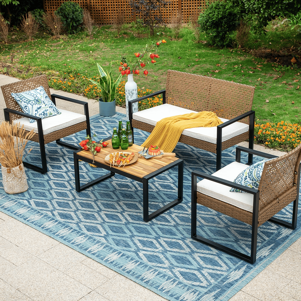 Outdoor patio set with two armchairs, a loveseat, and a wooden coffee table on a blue patterned rug. Includes white cushions, decorative pillows, a yellow throw, and is accessorized with a plant, vase of flowers, drinks, and a cheese platter.