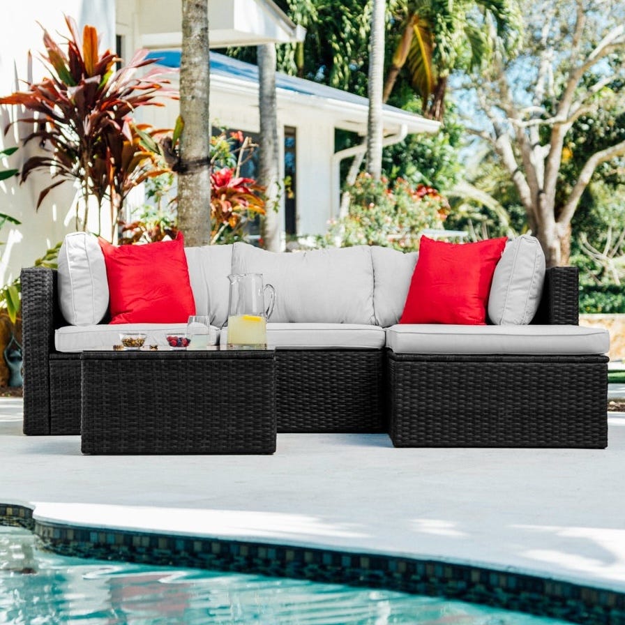 Outdoor patio set with a dark wicker sectional couch, white cushions, red pillows, and a matching coffee table, situated by a pool in a tropical garden setting.