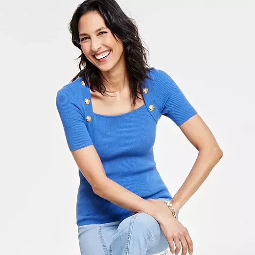 A blue square-neck sweater with short sleeves and gold-button embellishments on the shoulders. The wearer is paired with light jeans.